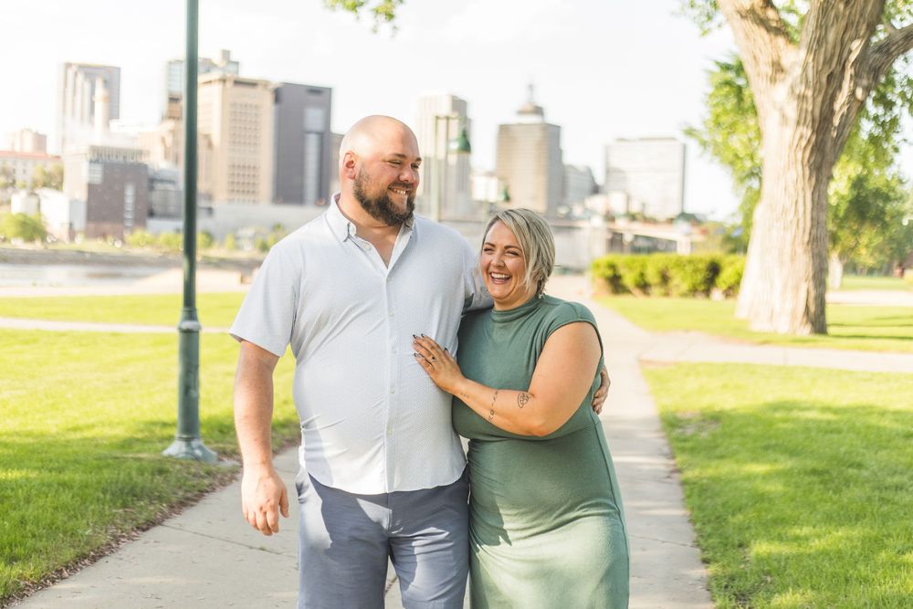 Smiling couple walking together on a sunny park path with a city skyline in the background.
