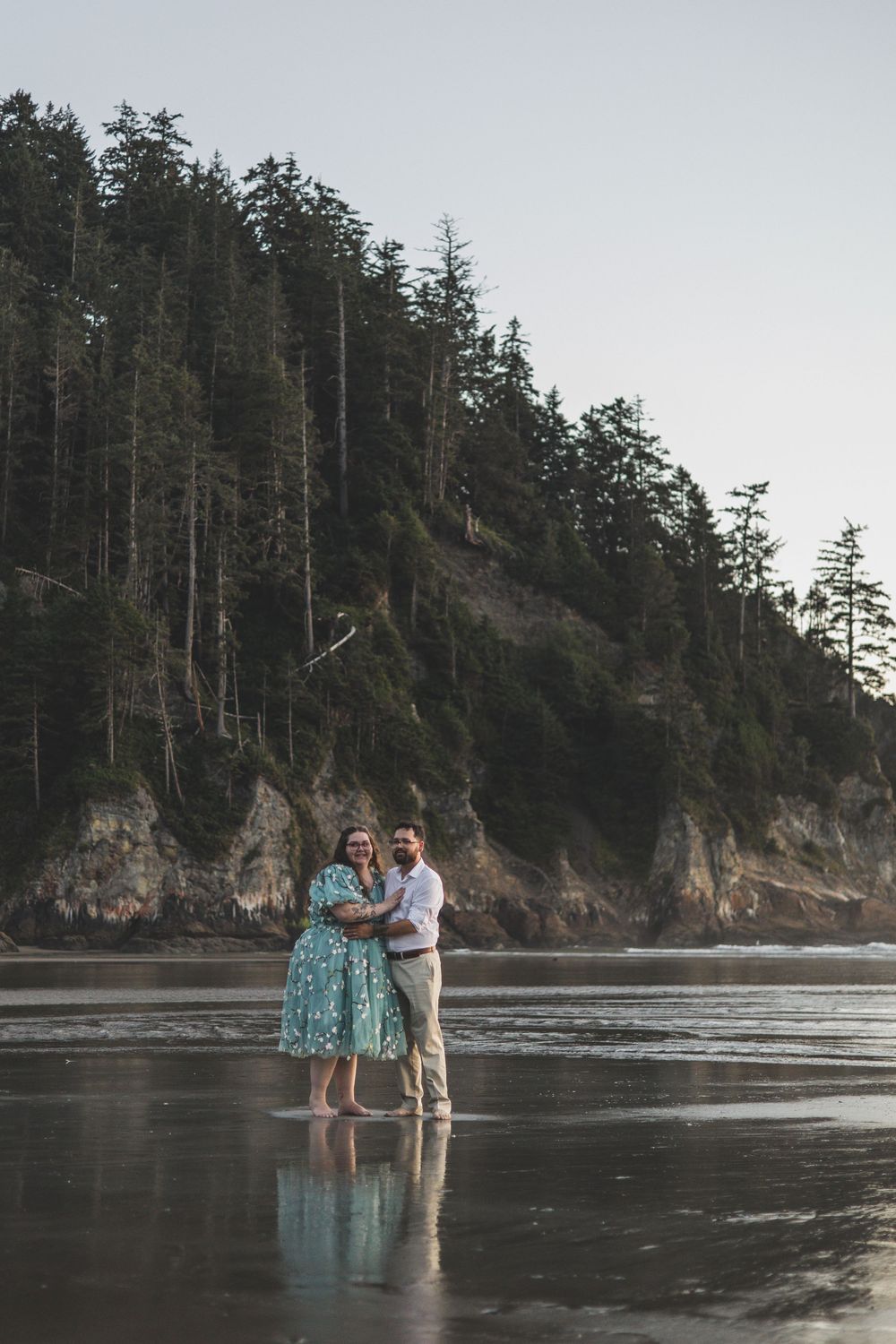 Couple embracing on a reflective beach with rocky cliffs and evergreen forest backdrop at dusk.