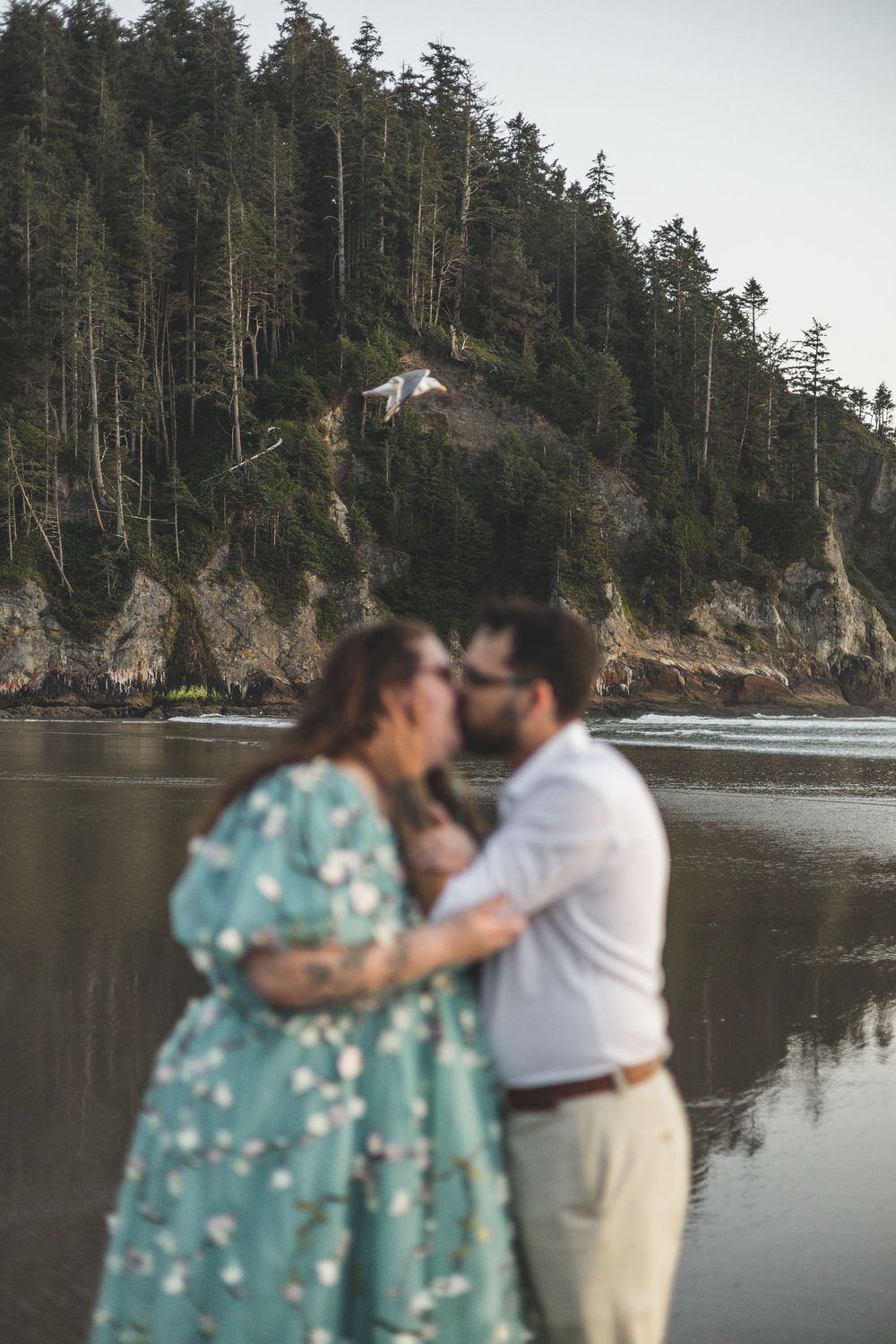 Couple sharing a kiss on a rocky beach with a forested cliff and calm water in the background.