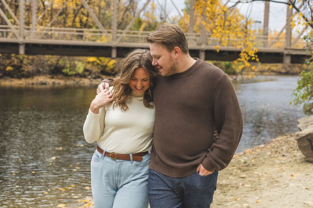 Couple smiling together by a river in autumn, wearing cozy sweaters with a wooden bridge in the background.