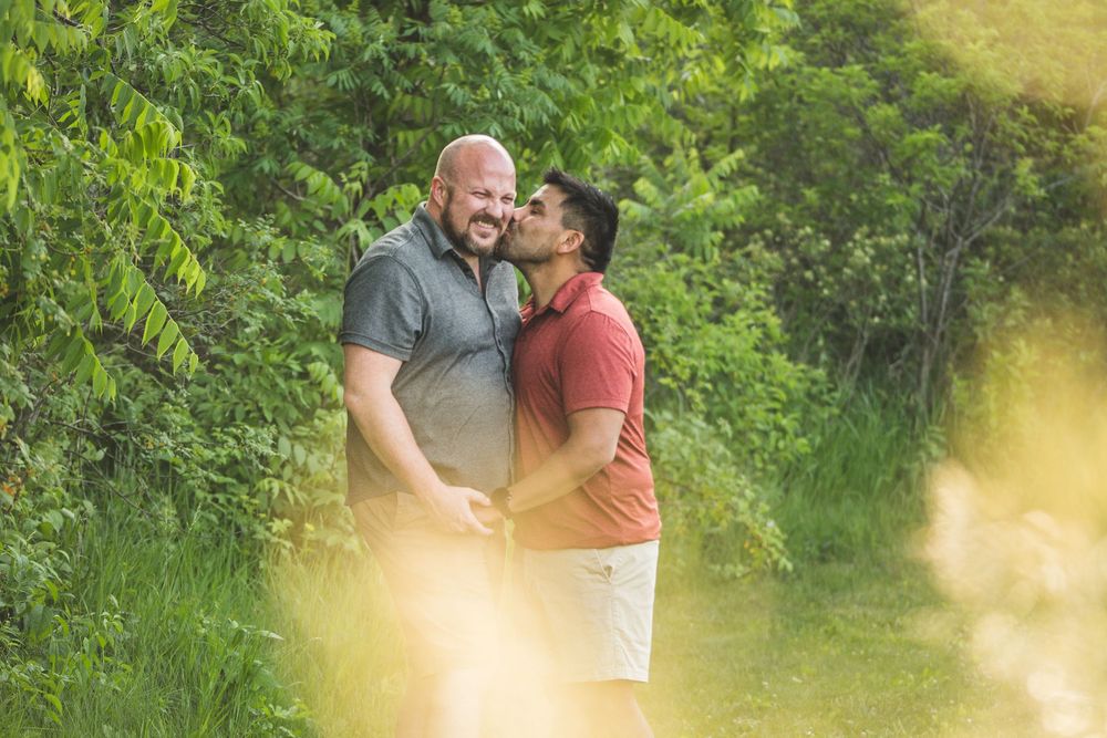Two men sharing a romantic moment outdoors, surrounded by lush green trees and foliage in a natural setting.