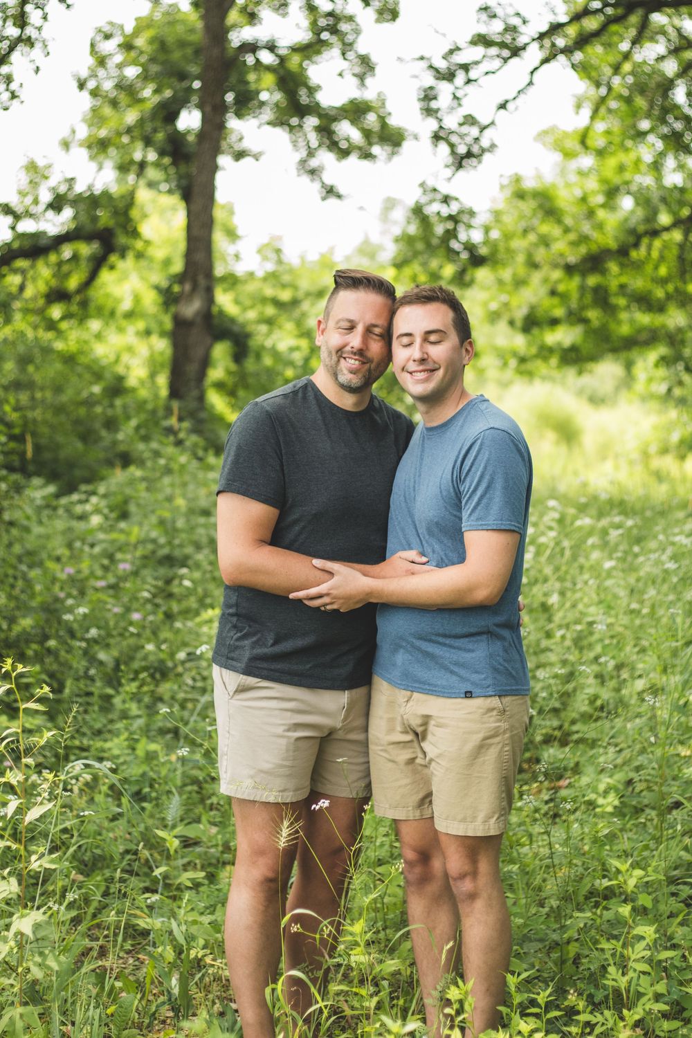 Two men embracing outdoors in a lush green forest setting, both wearing matching khaki shorts and casual t-shirts.