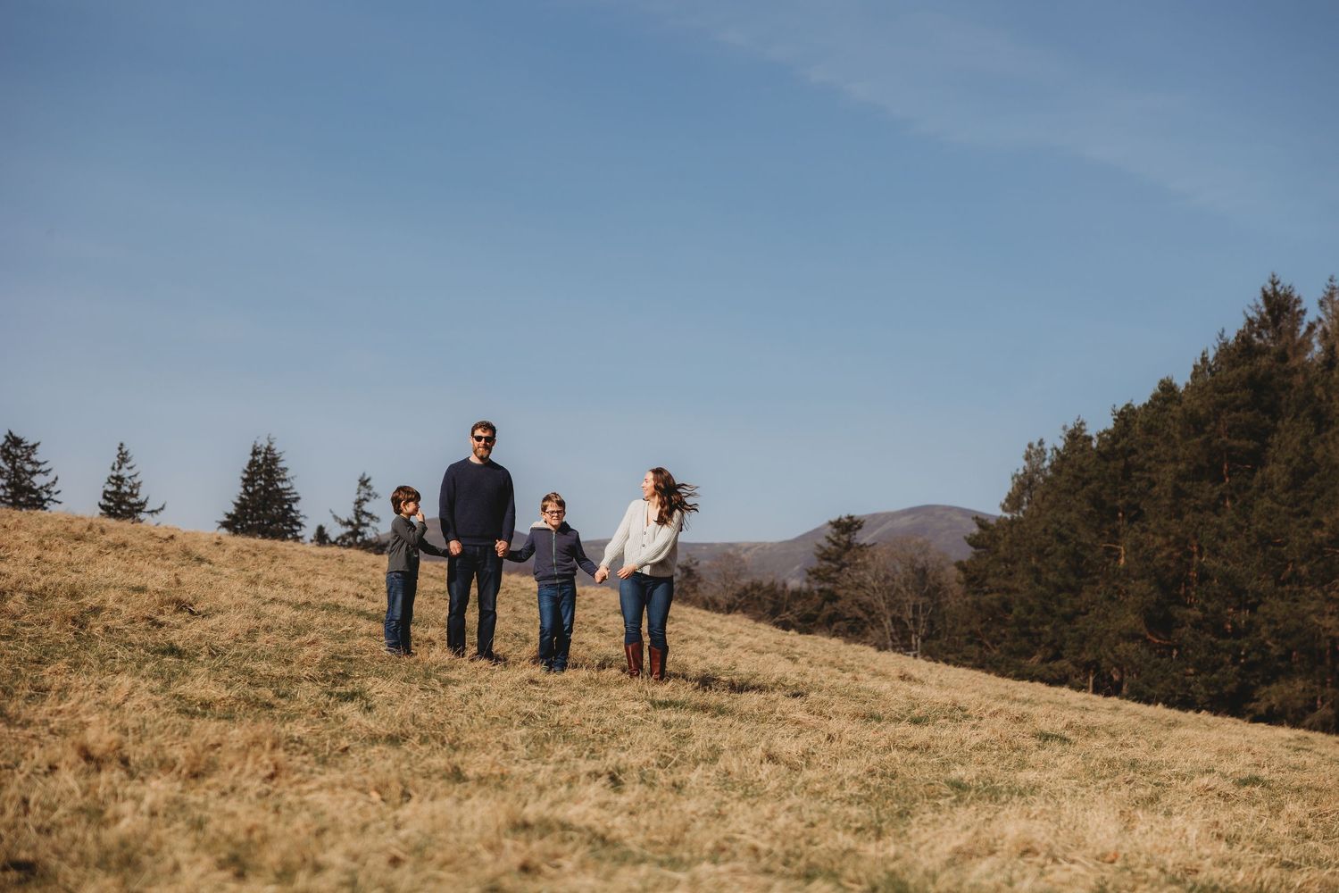 Family of four holding hands on a grassy hillside with pine trees and blue sky backdrop.