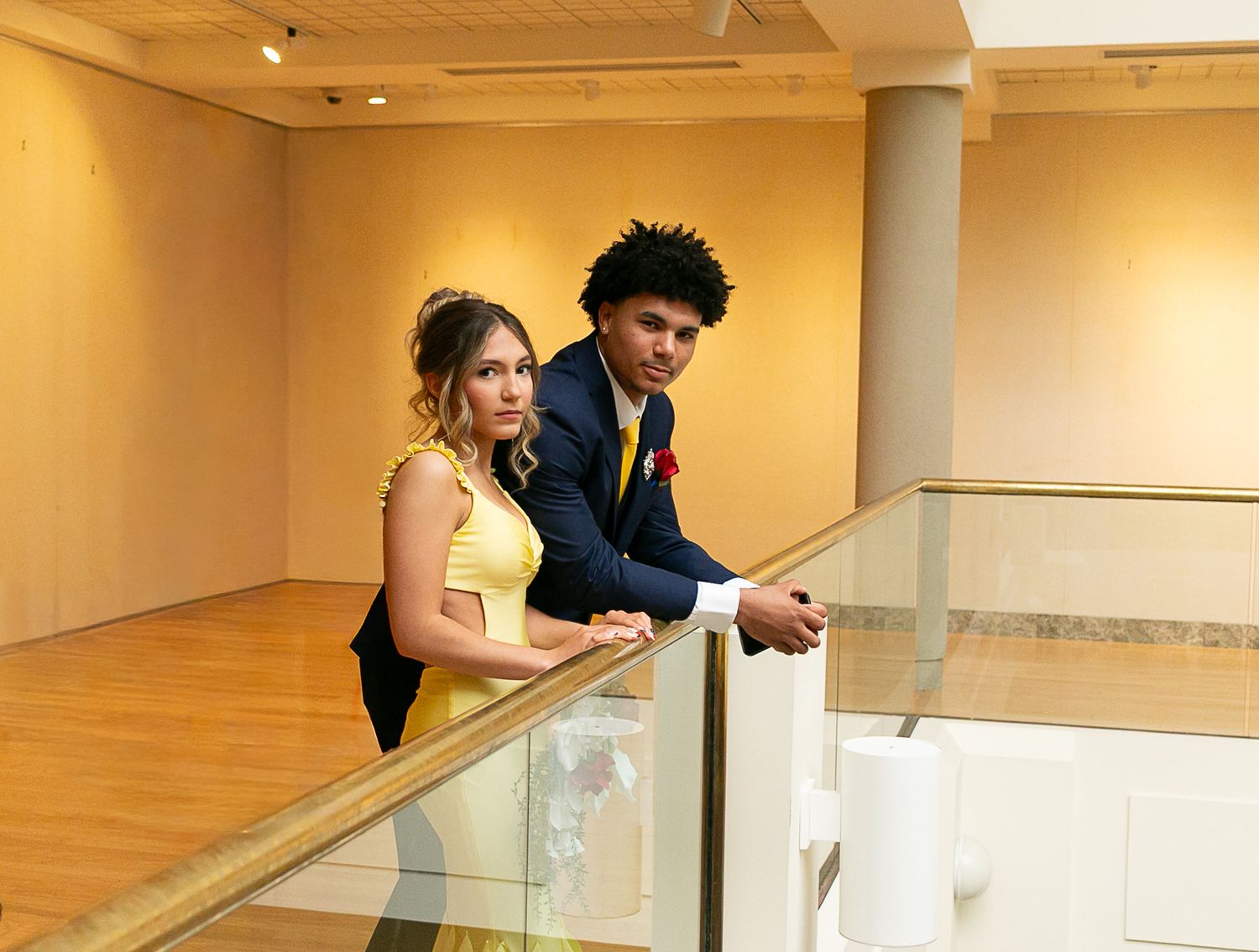 A young couple in formal prom attire pose elegantly by a glass railing in a modern gallery space.