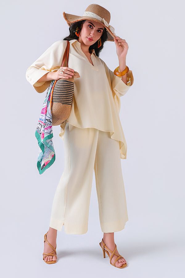 Woman in cream wide-leg pants and blouse, tan sun hat, strappy heels, carrying a woven bag with colorful scarf.