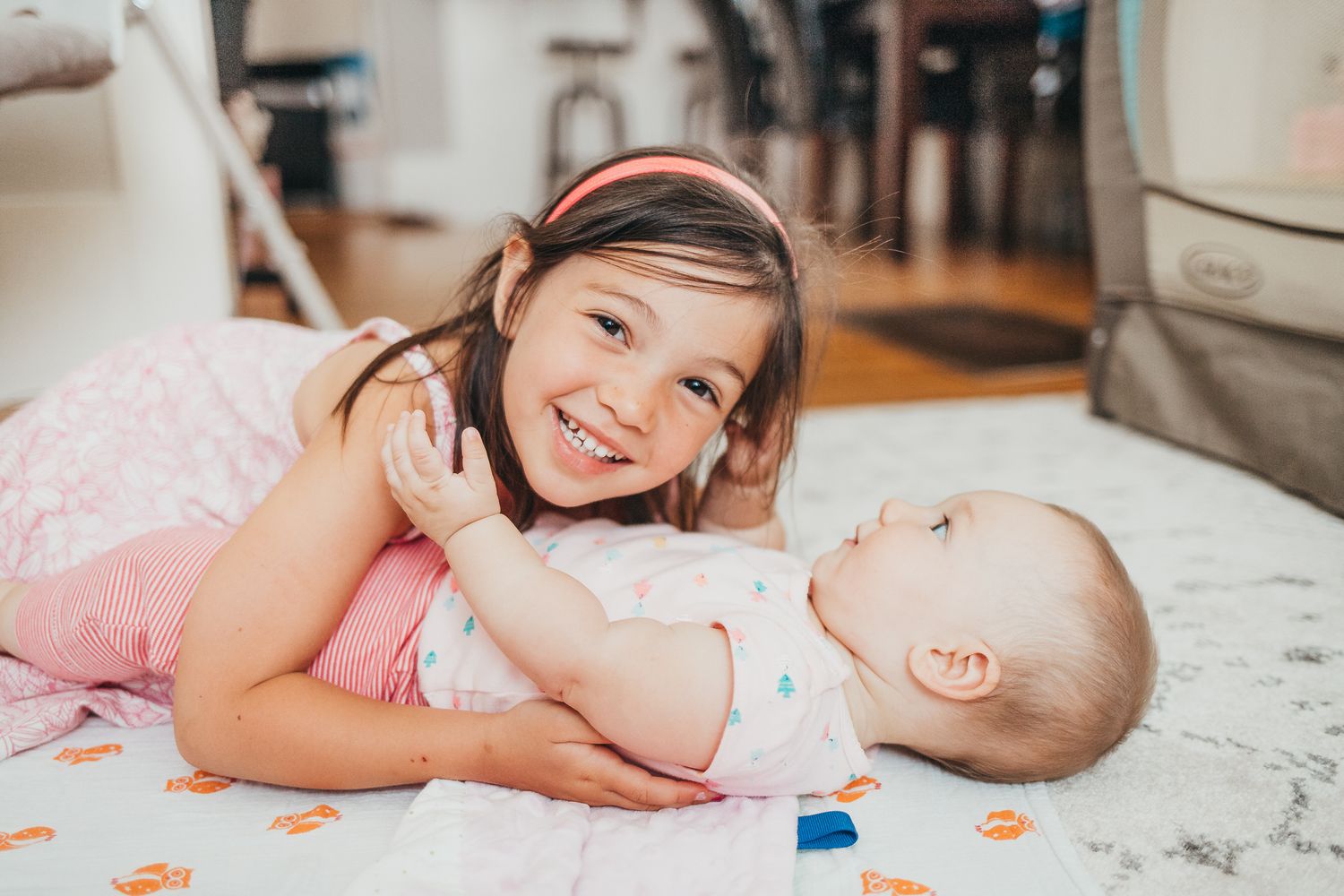 A smiling young girl in pink lies on the floor hugging a happy baby sibling in a bright kitchen.