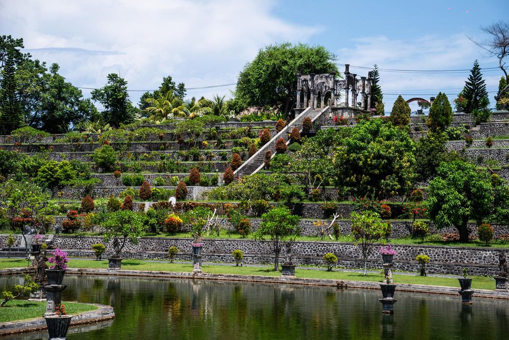 Terraced Balinese water palace garden with ornate stone sculptures, lush tropical plants, and a serene reflecting pond.