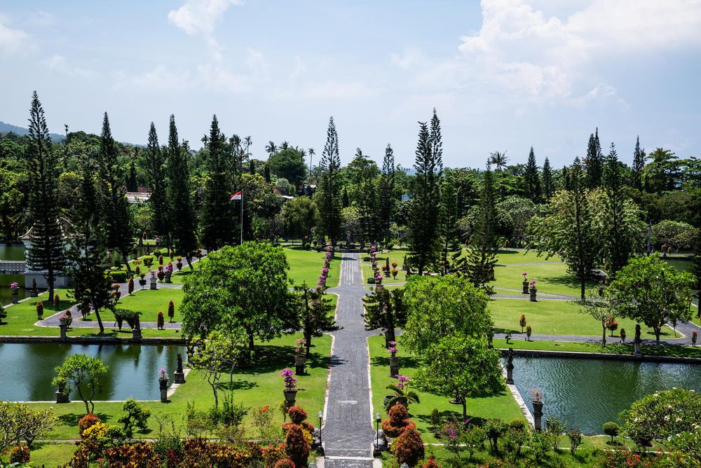 Lush tropical park with manicured gardens, tall cypress trees, and a central pathway surrounded by tranquil ponds.
