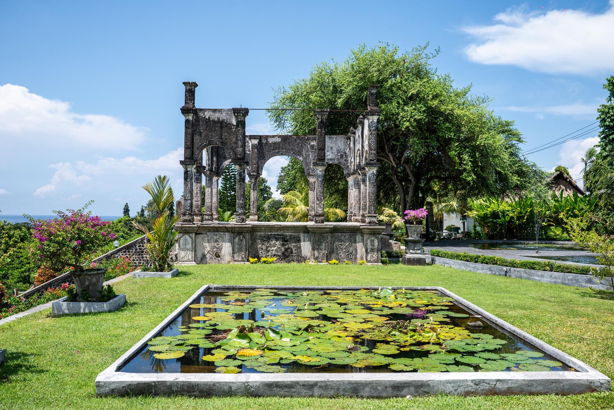 Ancient stone arch ruins reflected in a lily pond garden under a bright blue sky in Bali.
