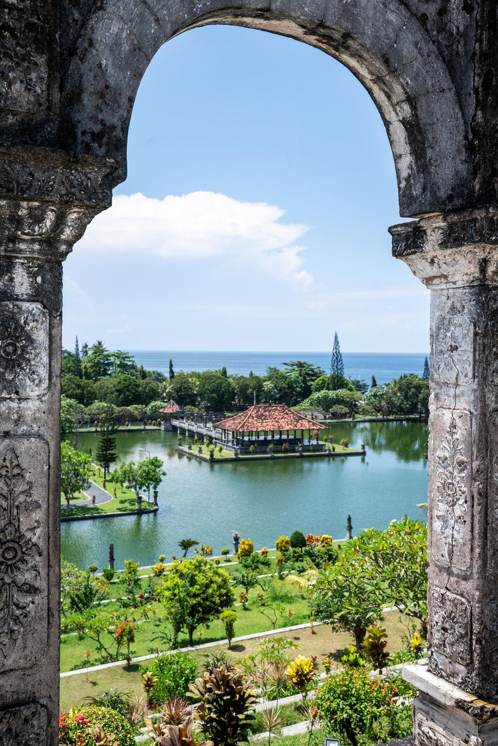 Stone arch frames a lush tropical water palace garden with pavilions and ocean views in Bali, Indonesia.