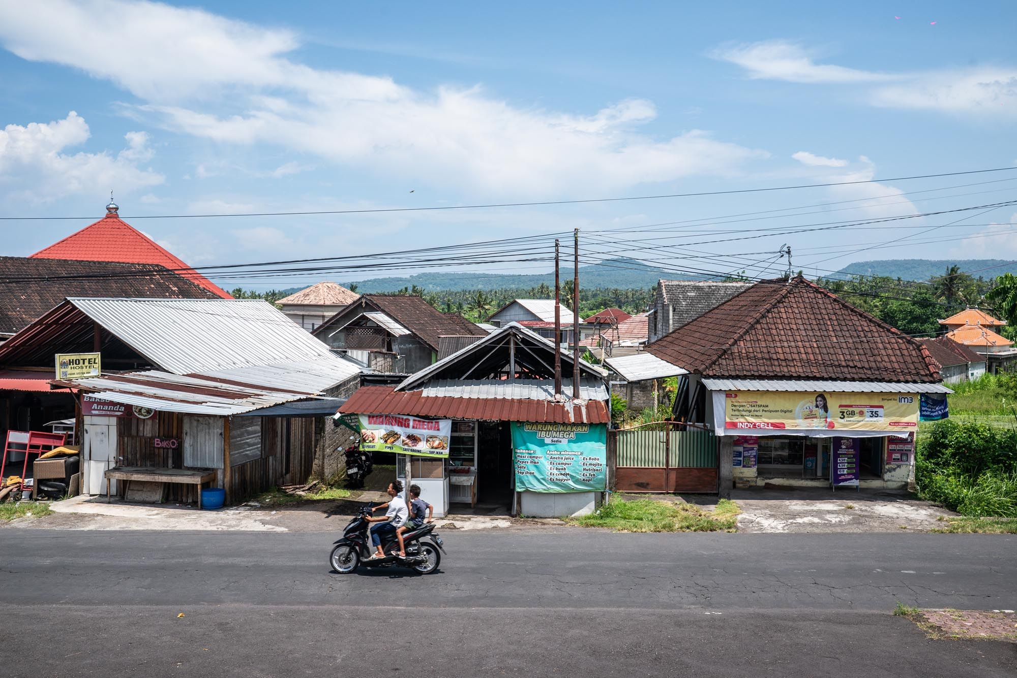 Motorcyclist rides past traditional Balinese buildings on a quiet street with lush green hills and blue sky in background.