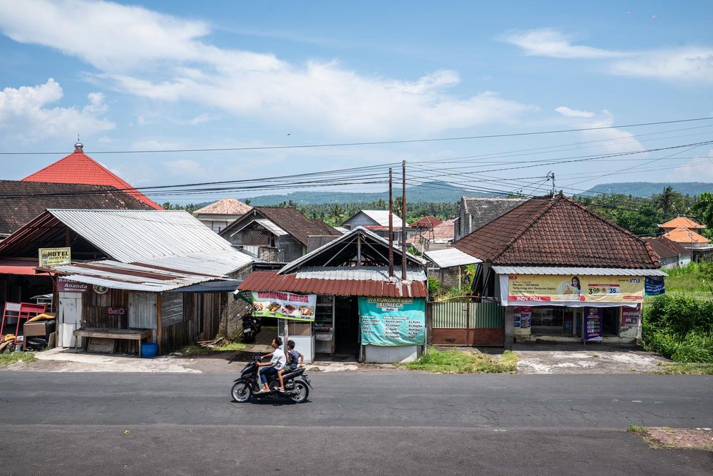 Motorcyclist rides past traditional Balinese buildings on a quiet street with lush green hills and blue sky in background.