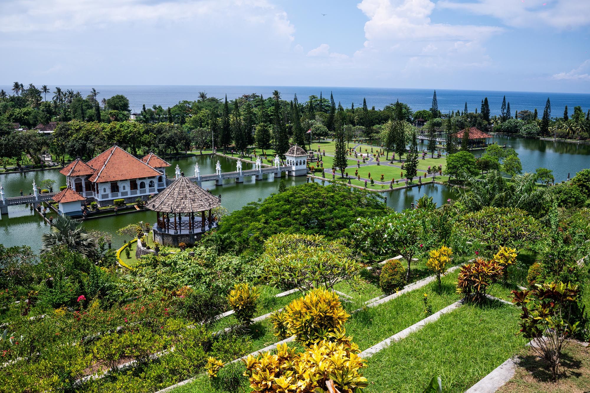 Aerial view of Taman Ujung water palace in Bali with tropical gardens, ponds, and ocean backdrop.