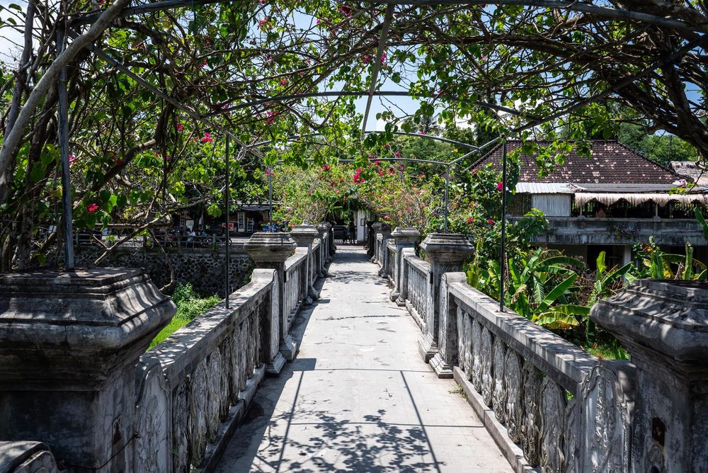 Stone pathway lined with ornate Balinese pillars and lush tropical trees leading through a serene garden.