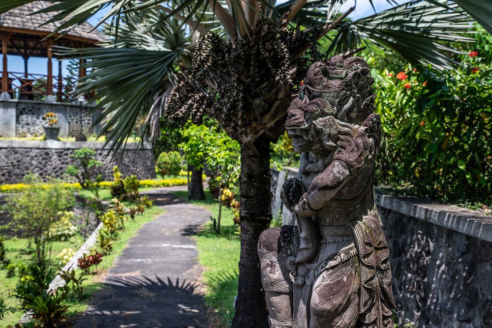 Ornate Balinese stone statue beside a palm tree in a lush tropical garden with a stone pathway.