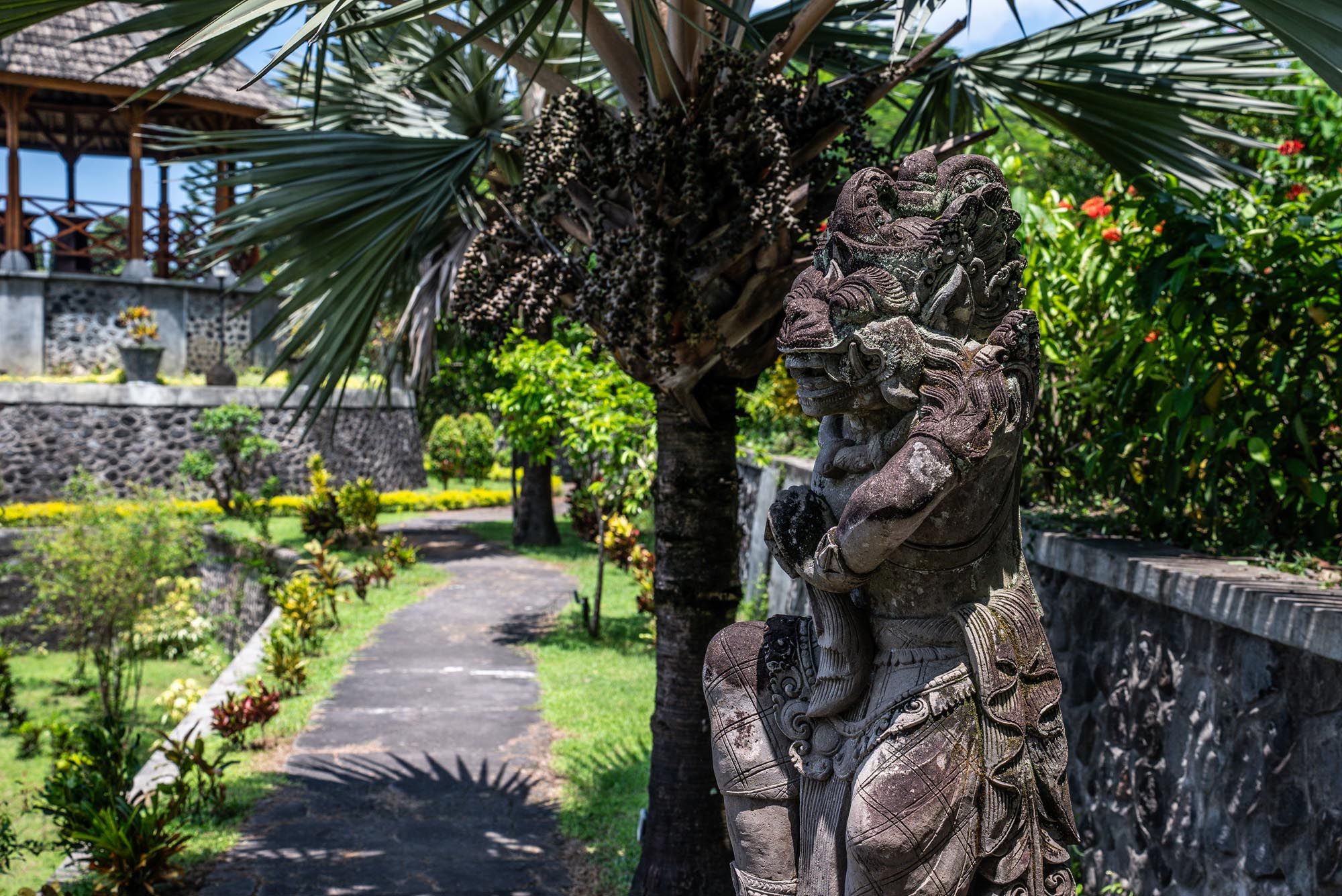 Ornate Balinese stone statue beside a palm tree in a lush tropical garden with a stone pathway.