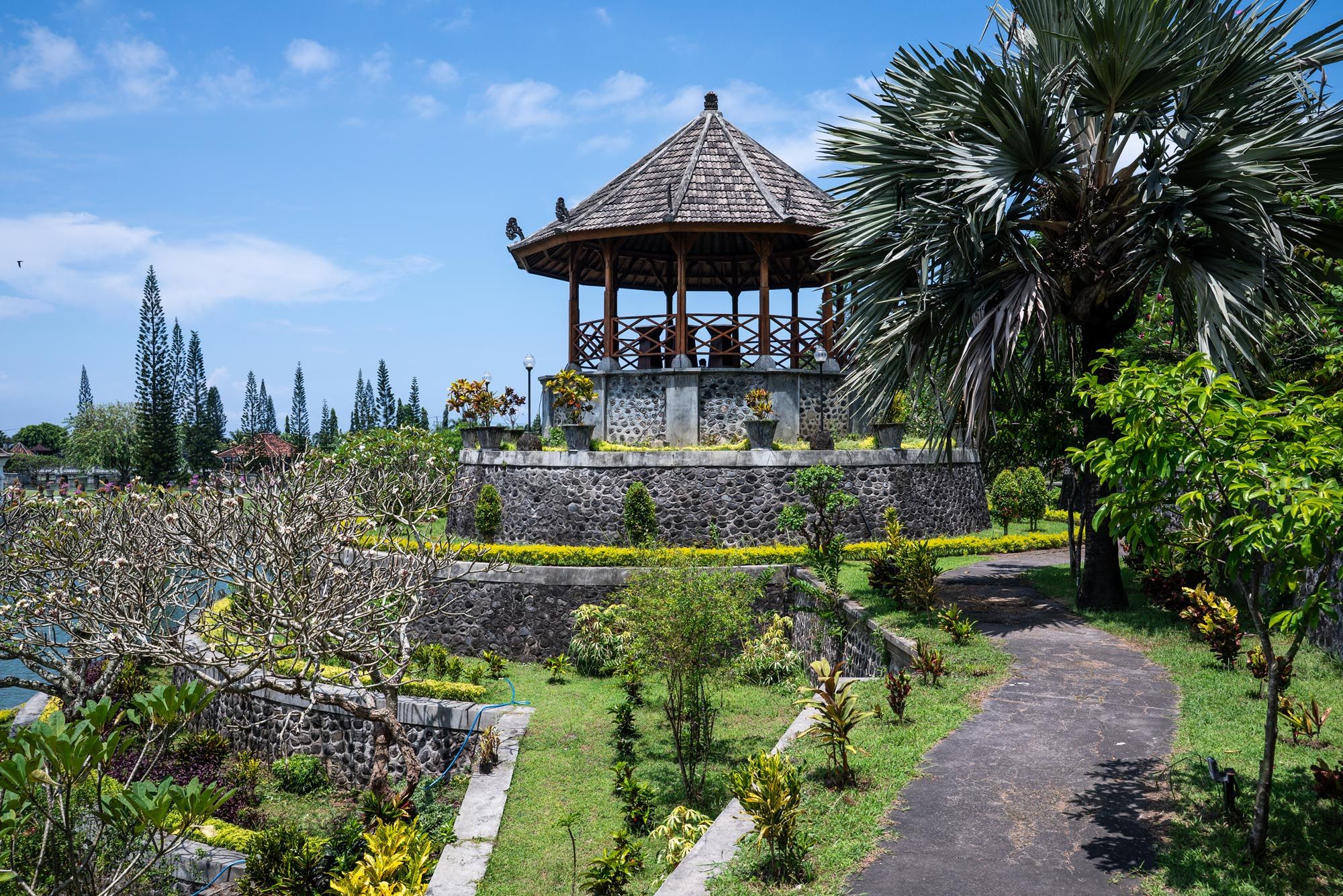 Traditional Balinese gazebo pavilion surrounded by tropical gardens and stone walls under a blue sky.