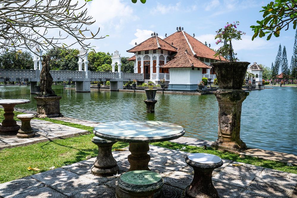 Balinese water palace with ornate stone tables, lush gardens, and a serene pond reflecting traditional architecture.