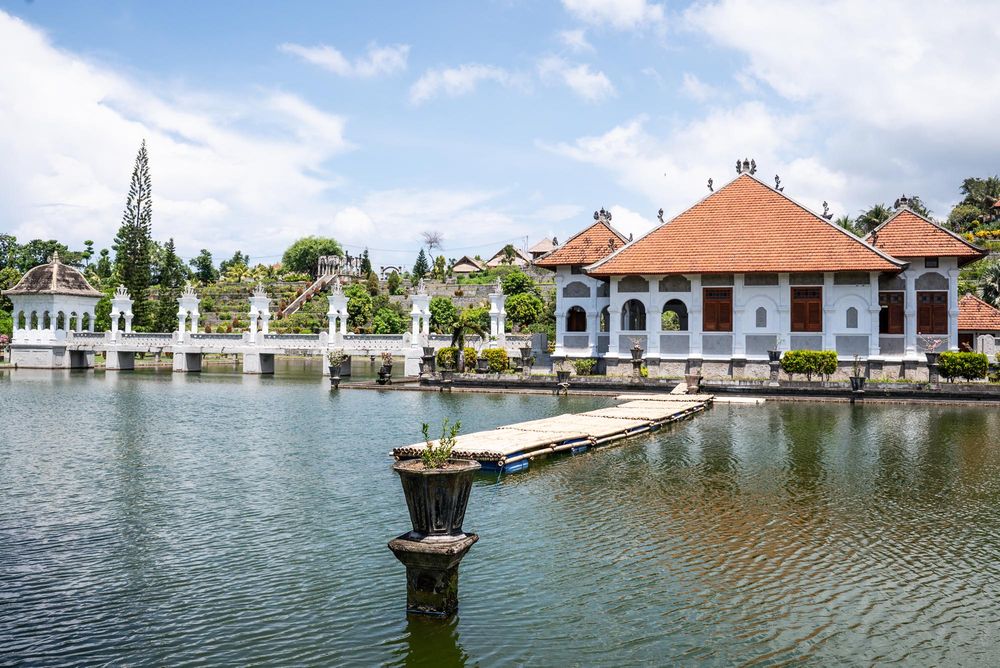Scenic Balinese water palace with white buildings, orange tile roofs, and serene lake surroundings under a partly cloudy sky.