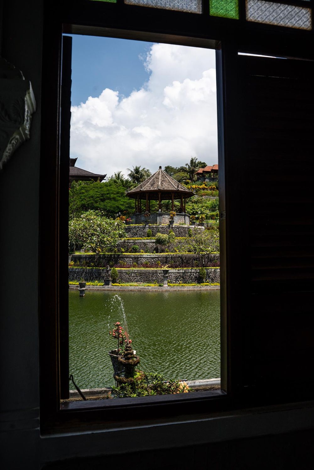 View through a dark window frame of a Balinese water palace with fountain, pond, and traditional pavilion.