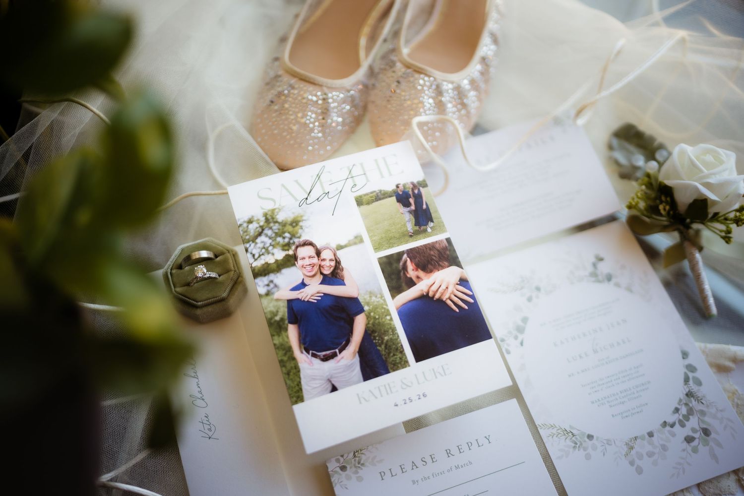 Wedding flat lay featuring invitation suite with couple photos, beaded bridal shoes, ring dish, and floral boutonniere on white background.