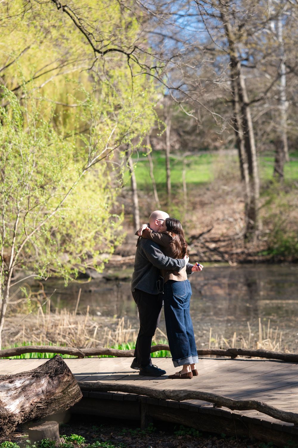 A couple embraces and kisses on a wooden bridge surrounded by spring trees and a peaceful pond.