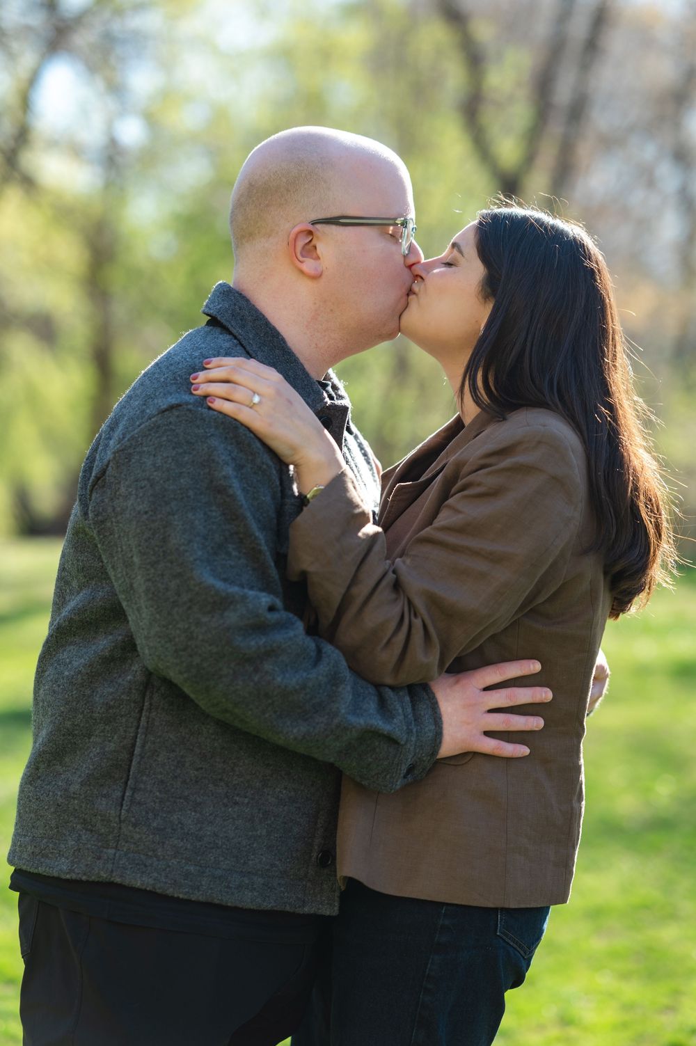 A couple shares a romantic kiss outdoors in a park during spring, man in gray jacket and woman in brown blazer embracing.
