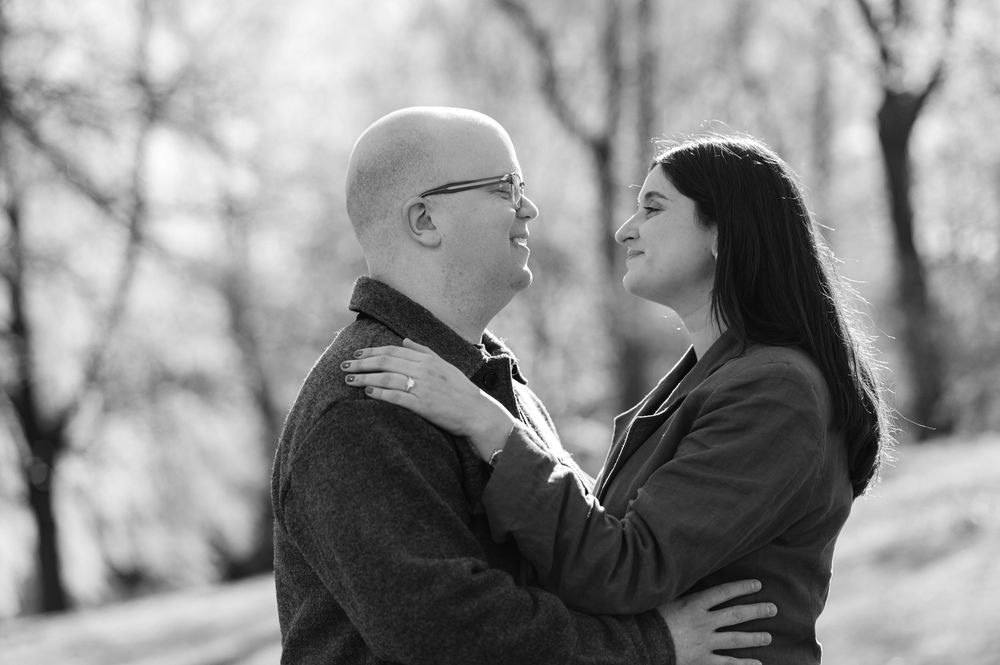 Black and white photo of a bald man and dark-haired woman embracing outdoors, gazing at each other romantically.