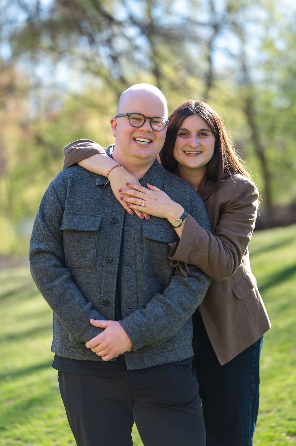 Smiling couple poses outdoors in a park during spring, woman embracing man from behind wearing glasses and gray jacket.