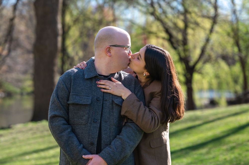 A couple shares a romantic kiss in a sunlit park with green trees and a pond in the background.