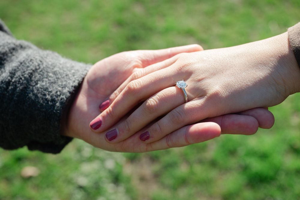 Close-up of a woman's hand wearing a diamond engagement ring held by her partner outdoors.