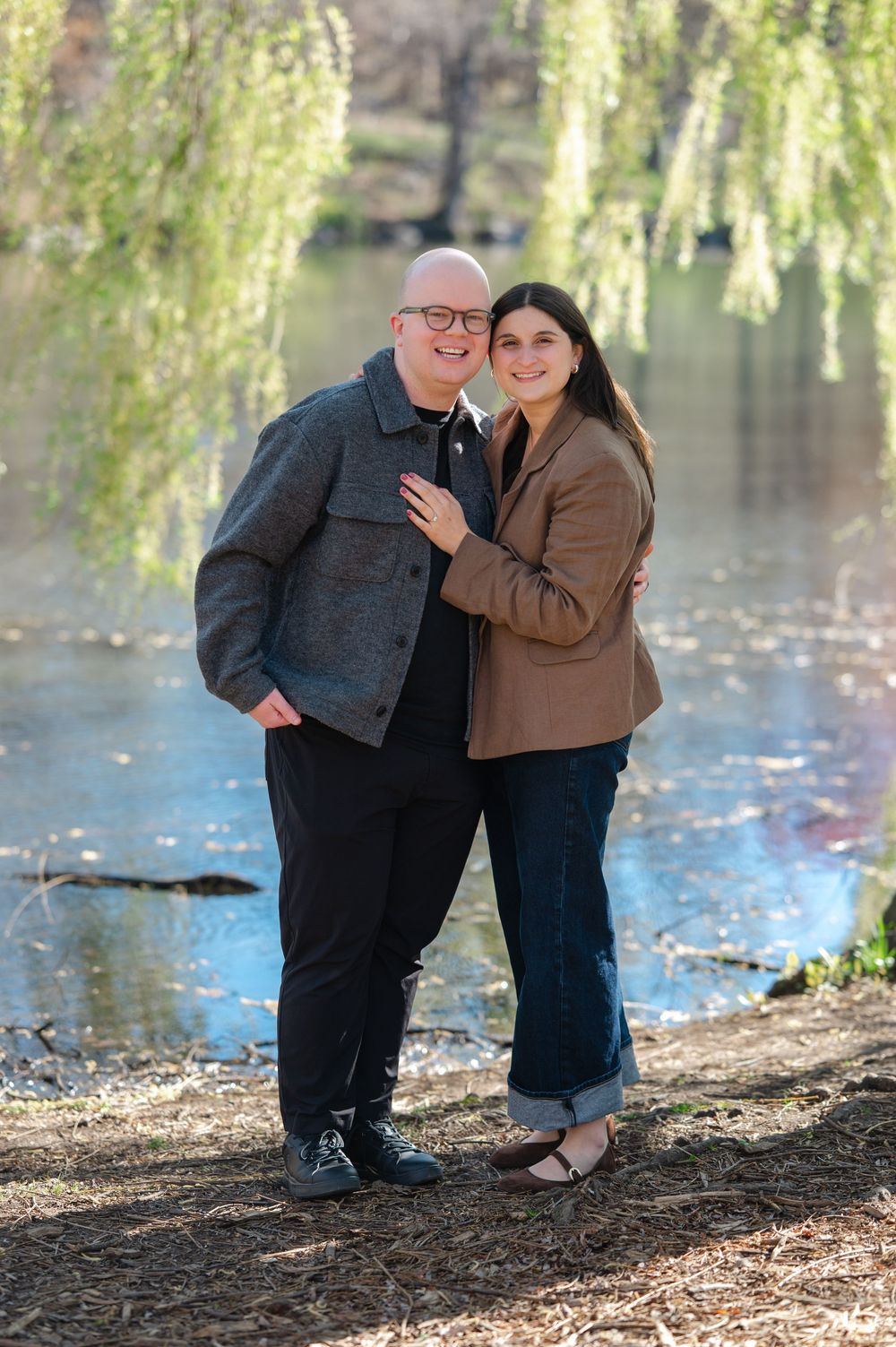 A happy couple poses together smiling near a weeping willow tree by a serene pond on a sunny day.