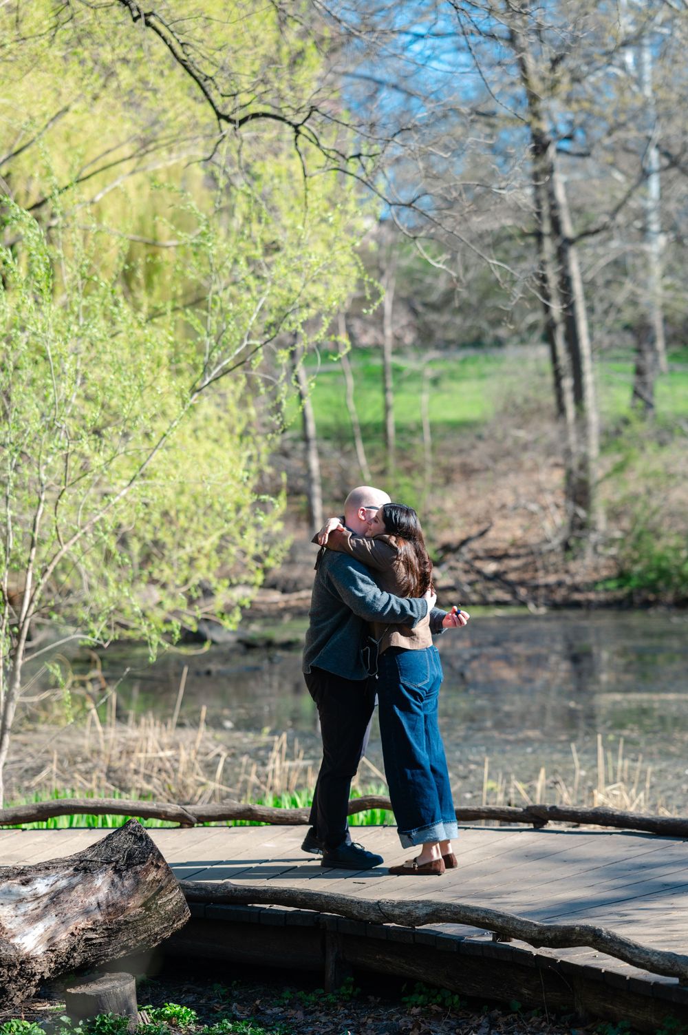 Couple embracing and kissing on a wooden bridge surrounded by trees and a pond in a lush park.