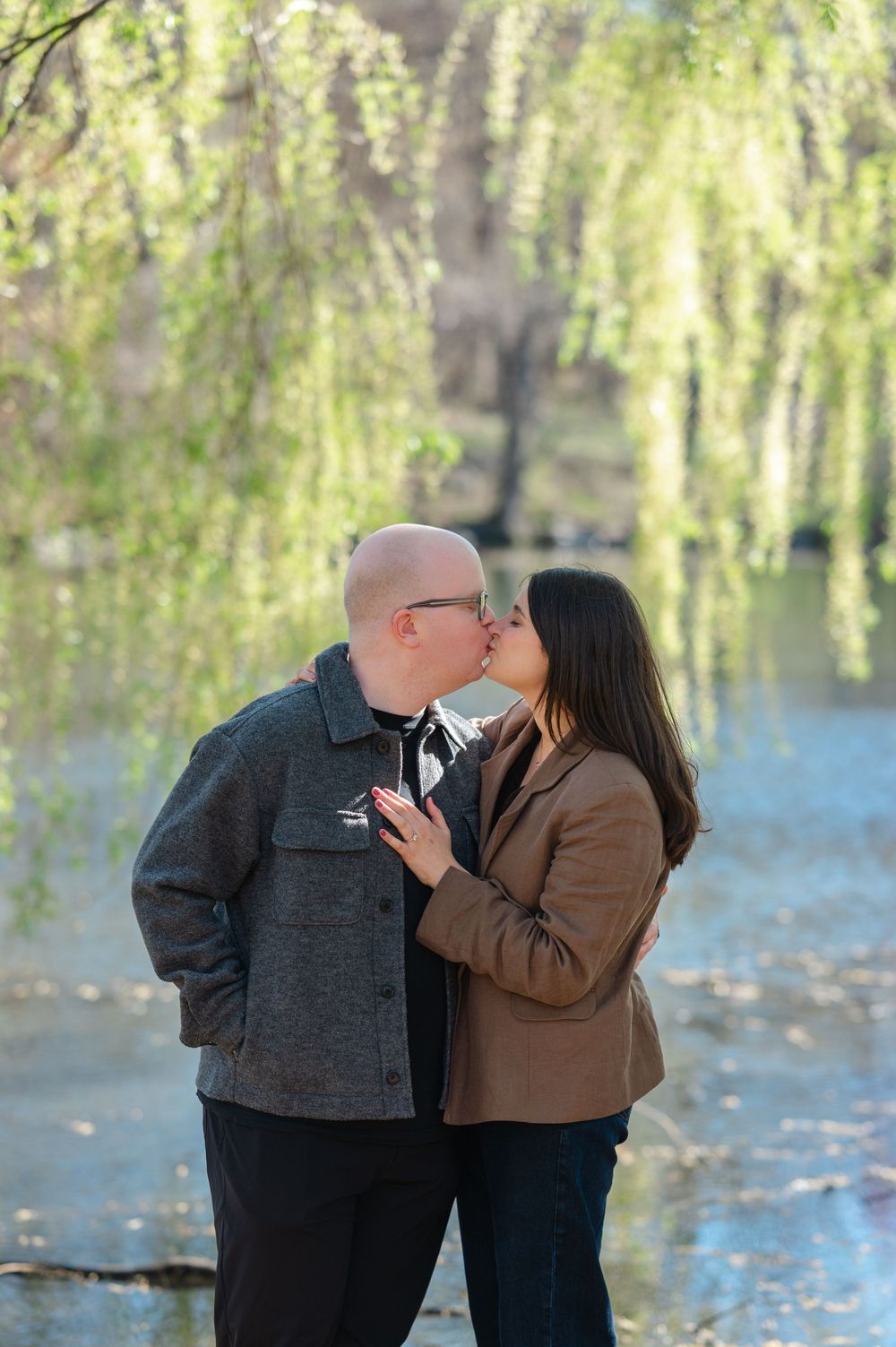Couple shares a romantic kiss near a weeping willow tree by a lake in a spring park setting.