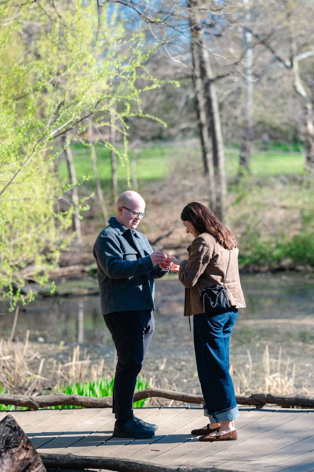 A man places an engagement ring on a woman's finger during a proposal on a scenic wooden dock by a pond.