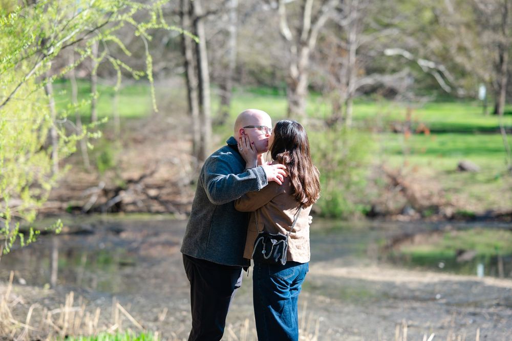 A couple shares a joyful kiss outdoors near a pond surrounded by spring trees after a marriage proposal.