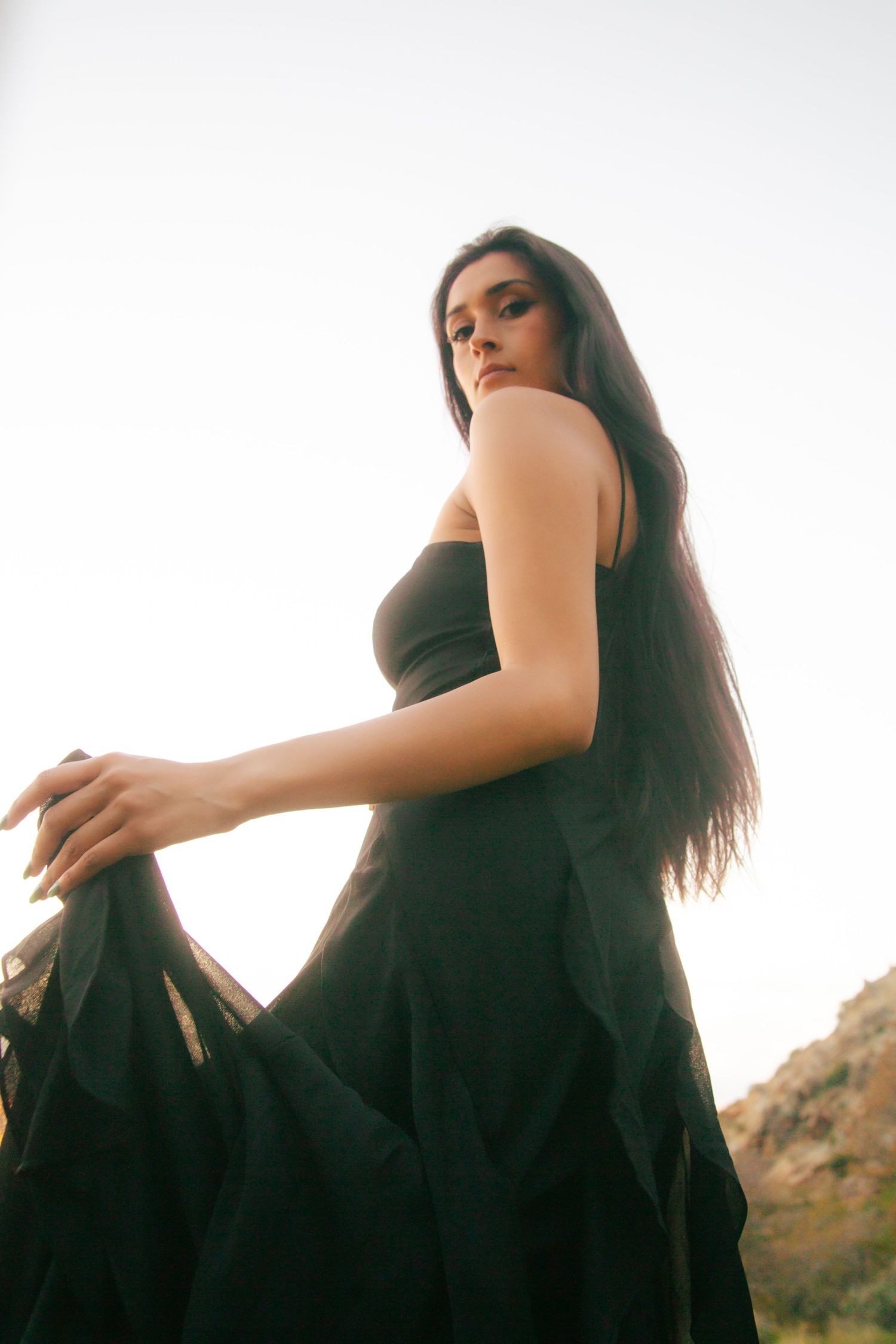 Low angle view of woman in black dress holding skirt with rocky mountain background at dusk.