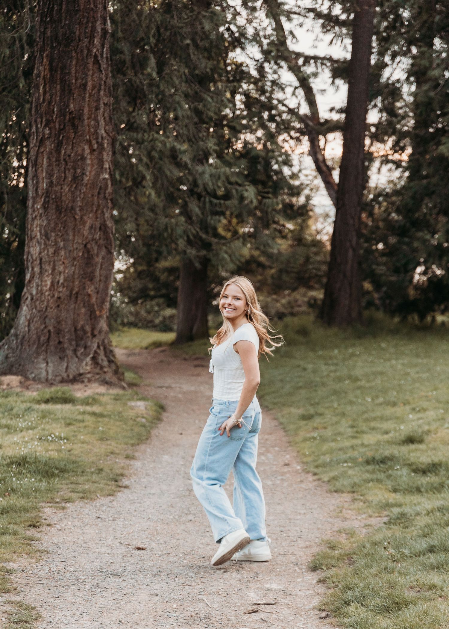 Young blonde woman smiling in light blue jeans and white top on a park path surrounded by tall trees.