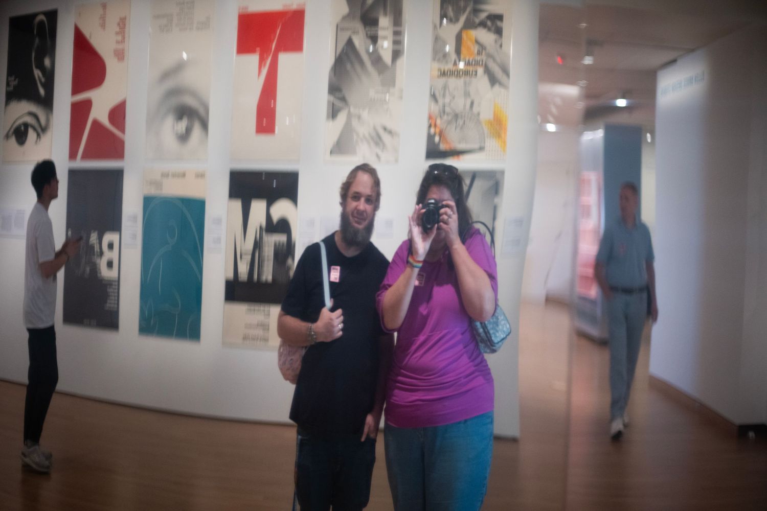 Two visitors pose for a photo in a modern art gallery with colorful graphic design posters displayed on the wall behind them.