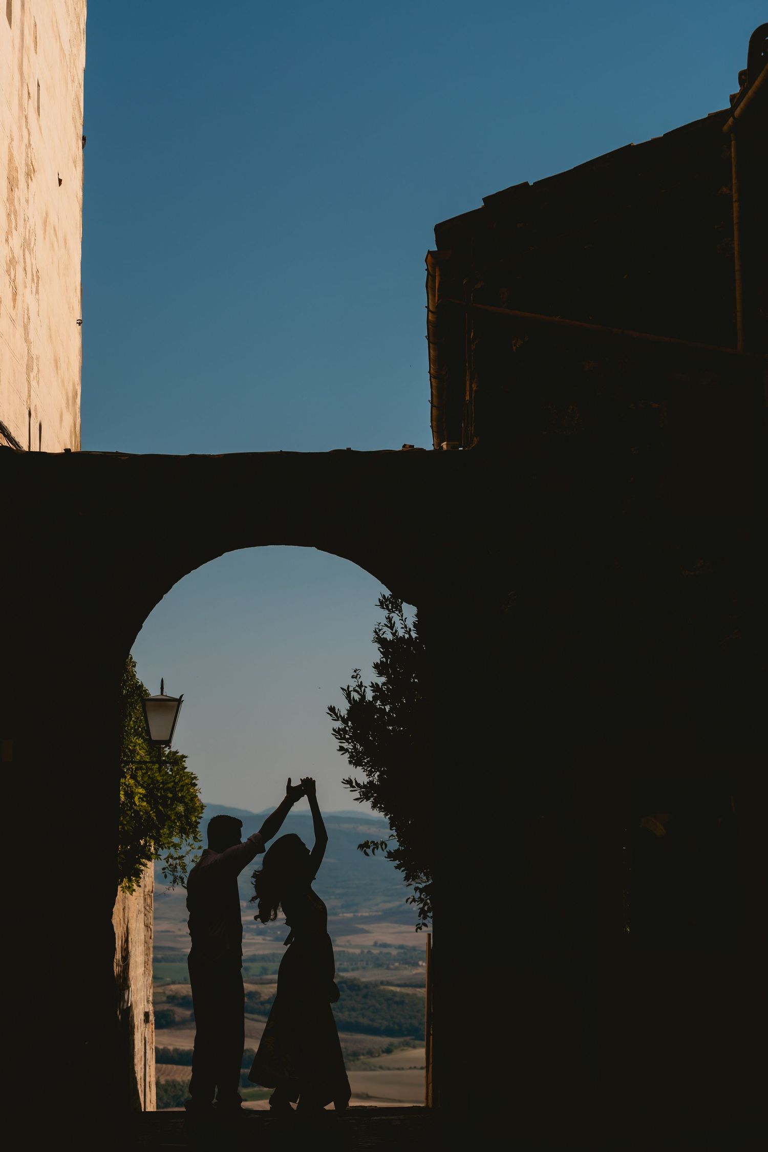 Romantic silhouette of couple facing each other under stone arch with scenic valley view behind them during an elopement in Pienza