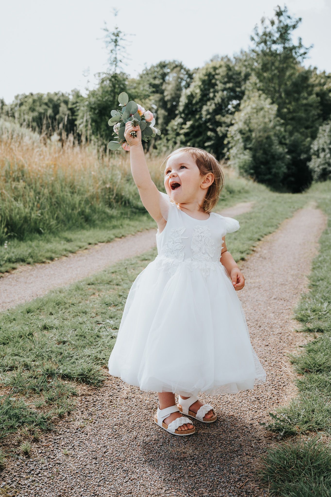 Joyful flower girl in white dress plays on a nature path surrounded by greenery.