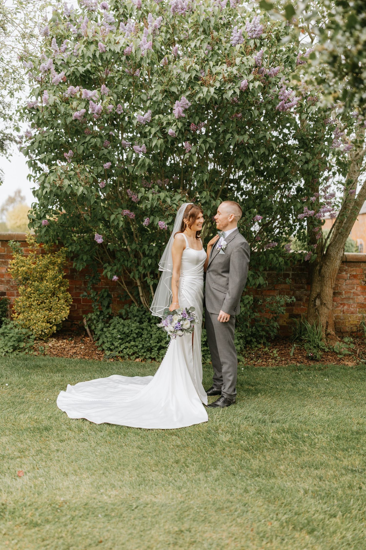 Bride and groom stand together on green lawn before blooming lilac tree, she in white gown, he in gray suit.