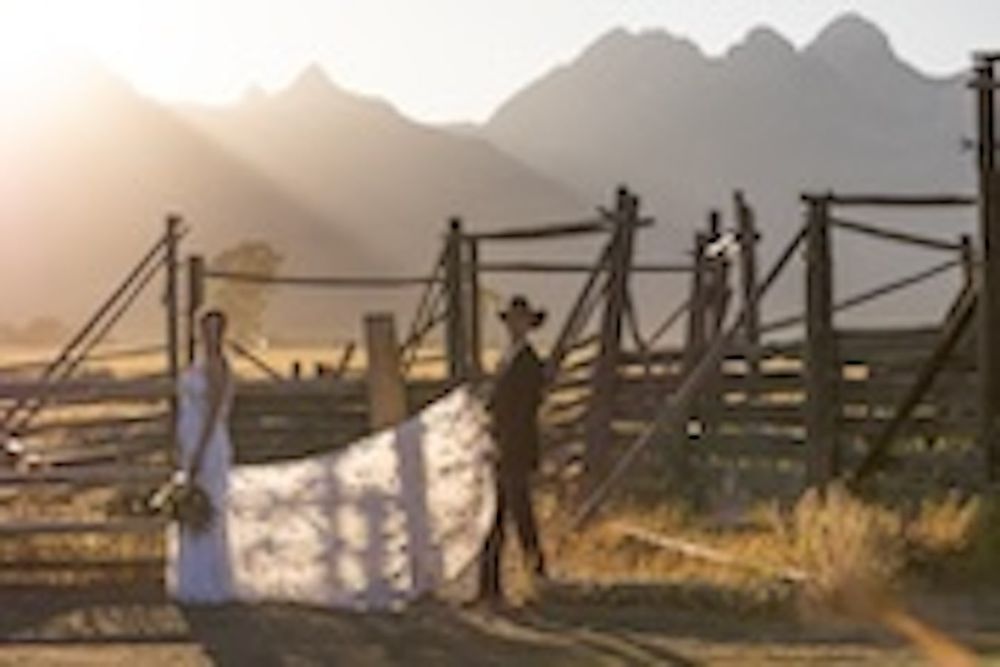 Bride and groom standing at rustic wooden gate with dramatic mountain backdrop at golden hour sunset.