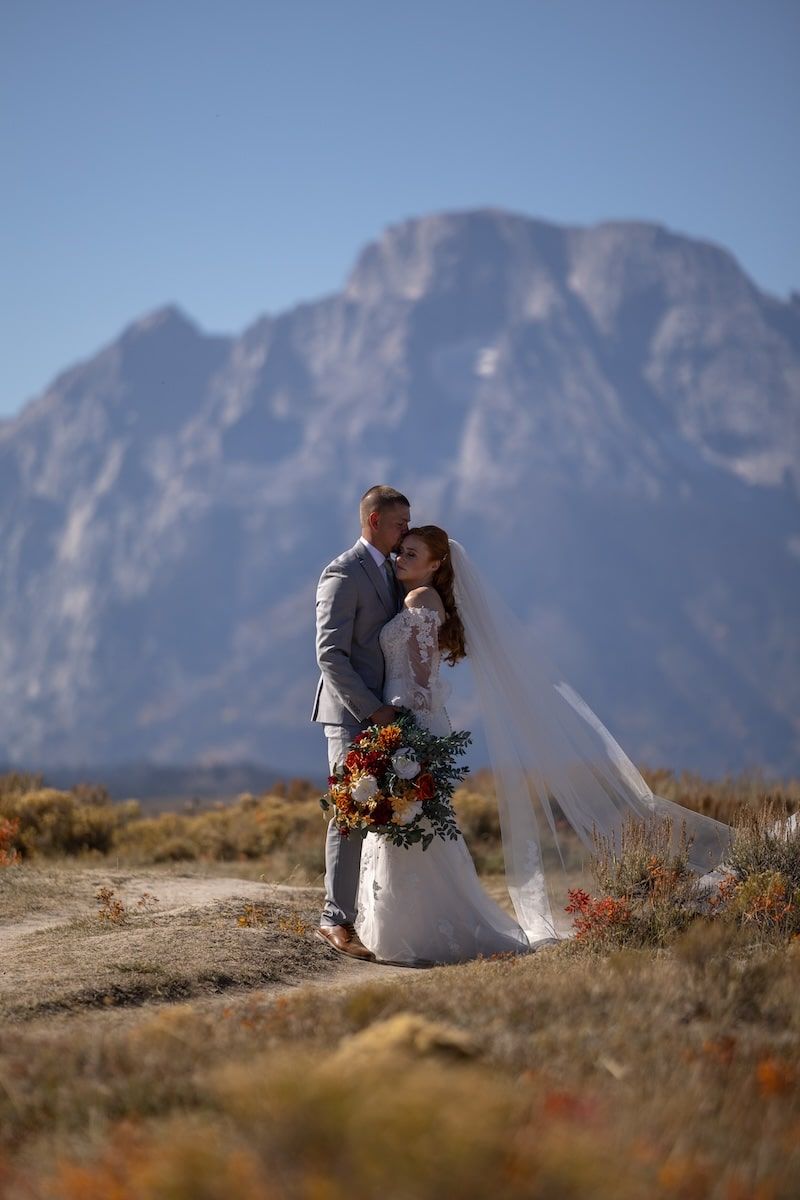 Bride and groom embrace on a mountain path, her veil flowing in the wind, with a stunning rocky peak backdrop.