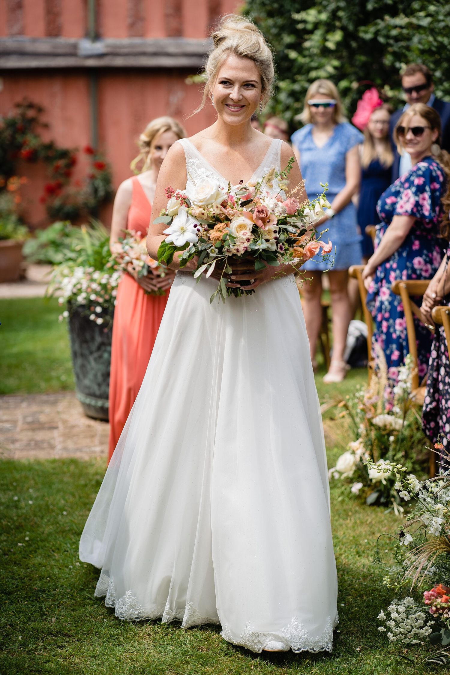 Smiling bride in white gown walks down outdoor aisle holding colorful bouquet, with guests and bridesmaid in coral dress behind her.