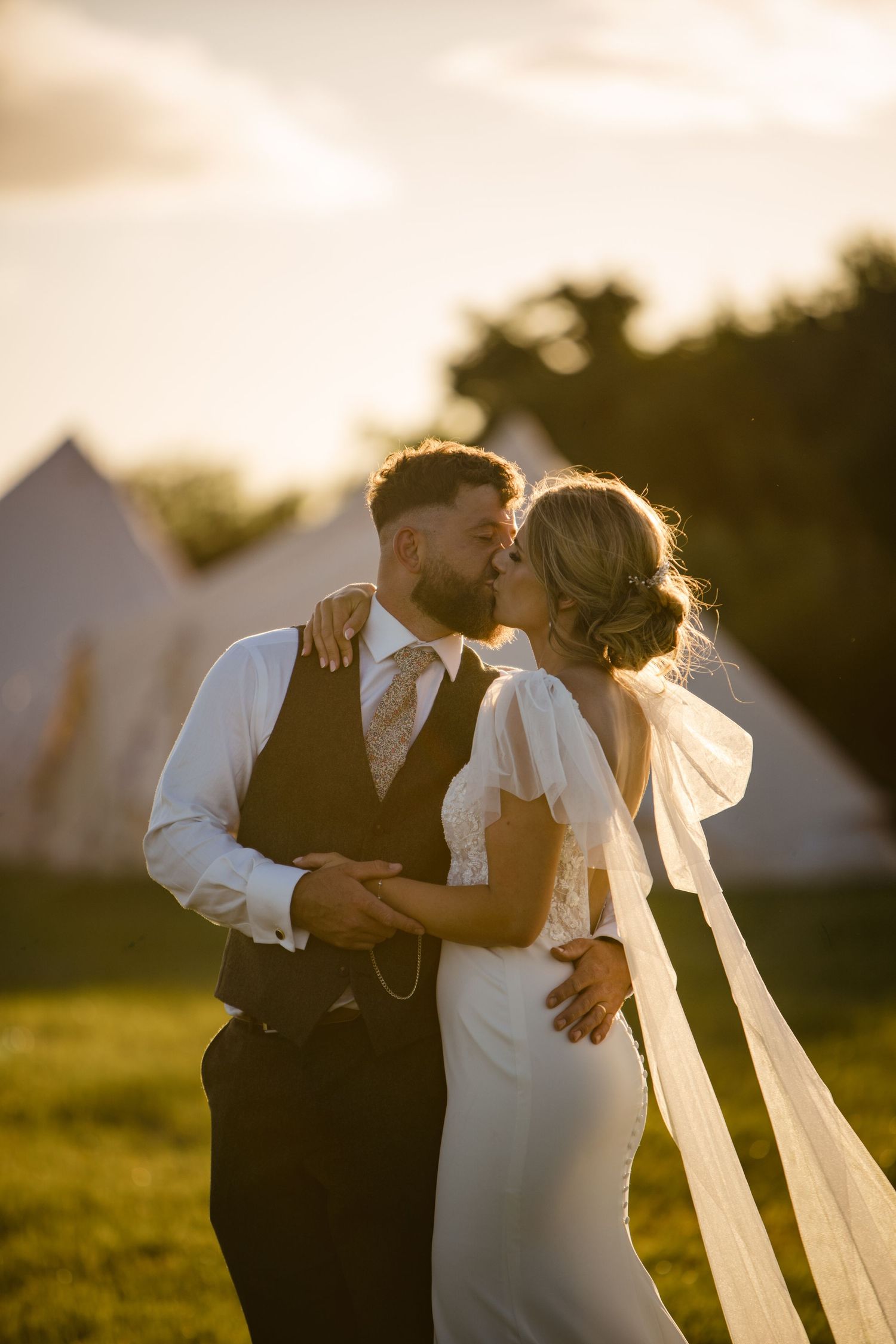 A bride and groom share a romantic kiss at golden hour, with tipi wedding tents in the background.