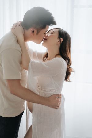 A romantic couple in white clothing embraces intimately near a bright window with sheer curtains.一對身穿白色服裝的浪漫情侶在明亮的窗戶旁親密擁抱，窗戶上掛著薄紗窗簾。