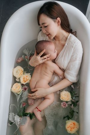 A tender milk bath photography session with mother and baby surrounded by floating peach roses and pink flowers.一組溫馨的牛奶浴攝影作品，母親和寶寶被漂浮的桃色玫瑰和粉