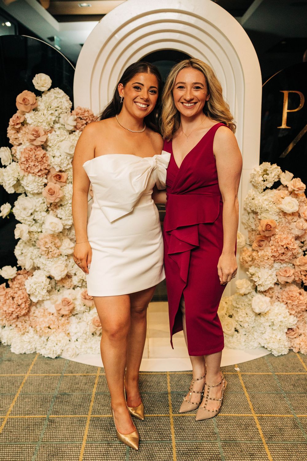 Two women posing together in front of floral arch, one in white mini dress and one in red midi dress.