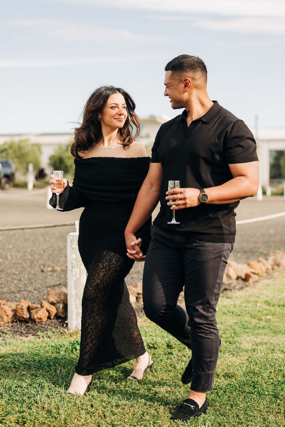 Elegant couple in black outfits holding champagne glasses while walking hand in hand at a winery.